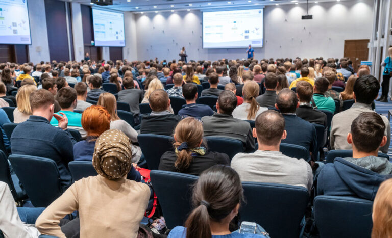 View from the back of a large conference room showing young professionals listening to a presenter during a session.