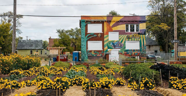 An urban farm plot of beds with crops and flowers sits in front of a building painted with an artistic mural, courtesy Michigan Urban Farming Initiative - MUFI